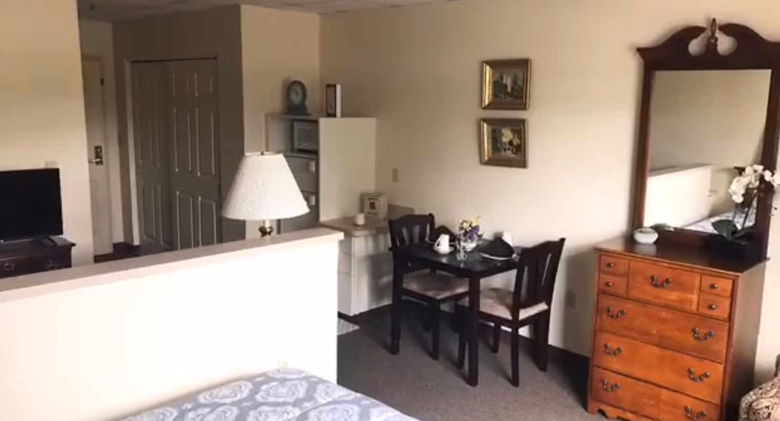 Interior view of a senior living facility room at Lehigh Commons showing a small dining table with two chairs, a wooden dresser with a mirror and decorative items, a bed partially visible in the foreground, and a lamp on a half wall separating the sleeping area from the dining space.