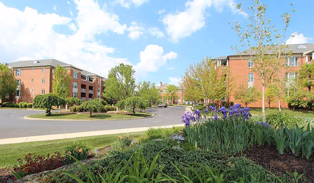 View of a senior living facility with red brick buildings, a circular driveway, and landscaped gardens featuring green shrubs and purple flowers under a partly cloudy sky.