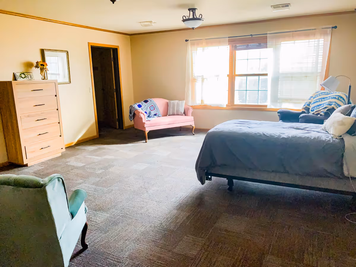 A spacious bedroom with a large window letting in natural light. The room features a bed with gray bedding, a pink loveseat with a crocheted blanket, a green armchair, a wooden dresser with a small vase of sunflowers and a framed picture above it, and a floor lamp next to the bed.