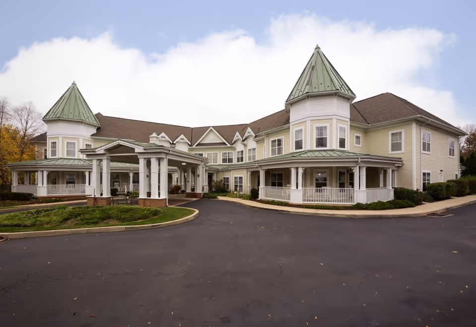 Exterior view of a large senior living facility building with two prominent turret-like structures topped with green roofs, a covered entrance, and a paved driveway. The building is light yellow with white trim and surrounded by some greenery and bushes under a partly cloudy sky.