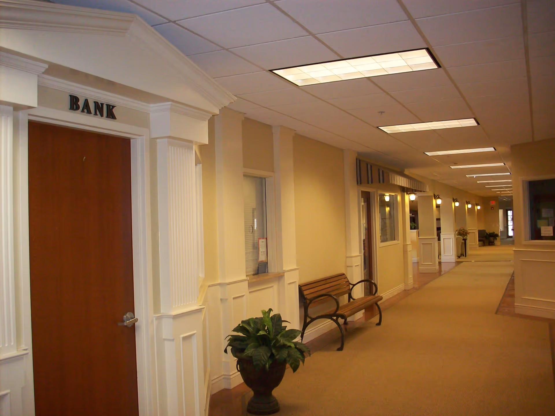 Interior hallway of a senior living facility with beige walls, carpeted floor, and ceiling lights. On the left side, there is a door labeled 'BANK' with white decorative molding around it. A wooden bench and a potted plant are placed along the wall. The hallway extends into the distance with multiple wall-mounted lights and windows on the right side.