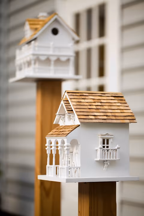 Two decorative white birdhouses with wooden shingle roofs mounted on wooden posts, positioned outdoors near a building with horizontal siding.