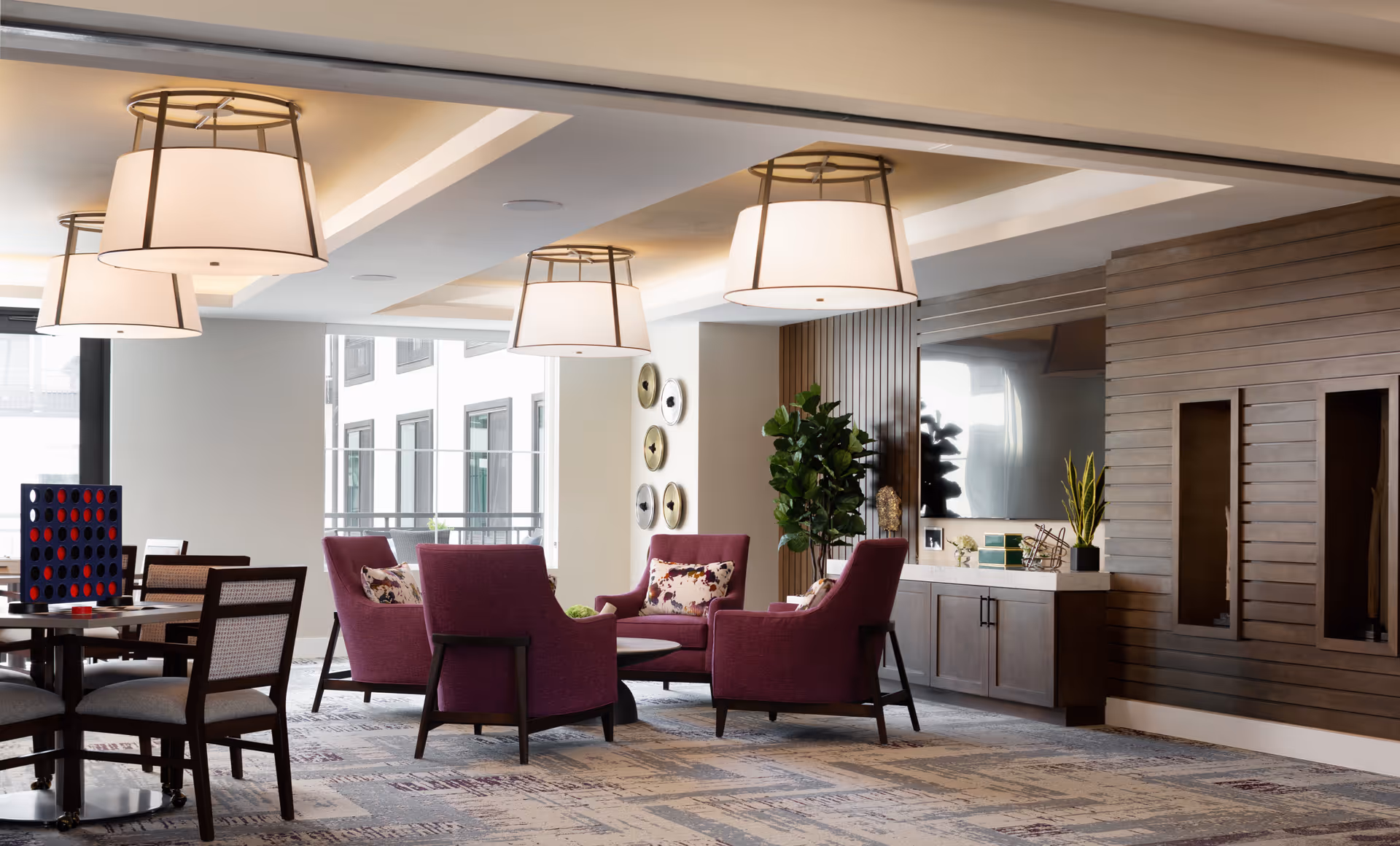 A modern senior living common area with four purple armchairs arranged around a circular coffee table. The room features large hanging ceiling lights, a patterned carpet, a flat-screen TV mounted on a wooden paneled wall, and decorative plants. There is also a table with chairs and a large Connect Four game near a window with a view of an adjacent building.