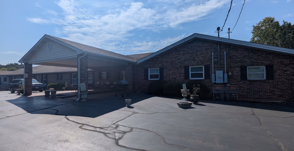 Exterior view of a single-story brick building with a covered entrance and a paved driveway. Several potted plants are placed near the building, and a few vehicles are parked nearby under a clear blue sky.