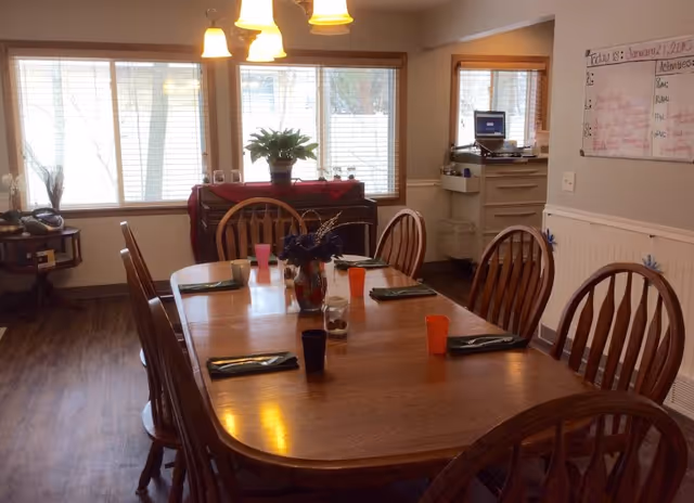 A wooden dining table with place settings and chairs in a bright room with large windows, a sideboard with a plant, and a whiteboard on the wall.