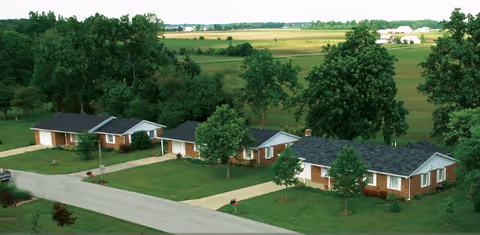 Aerial view of a senior living community with several single-story brick homes, each with a driveway and surrounded by well-maintained lawns and trees. In the background, there are expansive green fields and a few distant buildings under a clear sky.