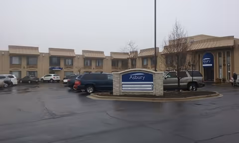 Exterior view of Asbury Court Senior Living Community building with a parking lot in front. Several cars are parked and a sign with the facility name is visible in the center of the image. The building is two stories with beige walls and awnings over the windows.