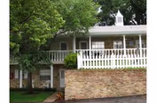 Exterior view of a brick and white-paneled building with a white fence and railing on the upper level, surrounded by trees and greenery.