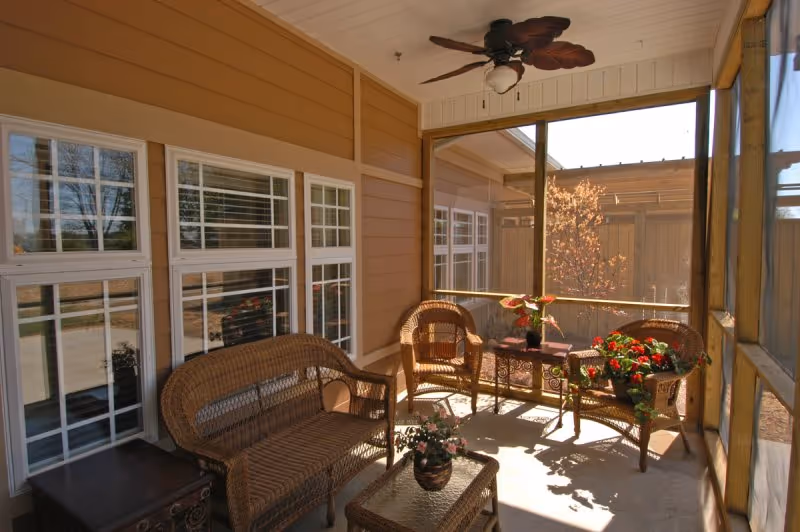 A screened-in porch area with wicker furniture including a loveseat, two chairs, and a glass-top coffee table. There are potted plants on the table and on the floor, a ceiling fan with leaf-shaped blades, and large windows looking outside.