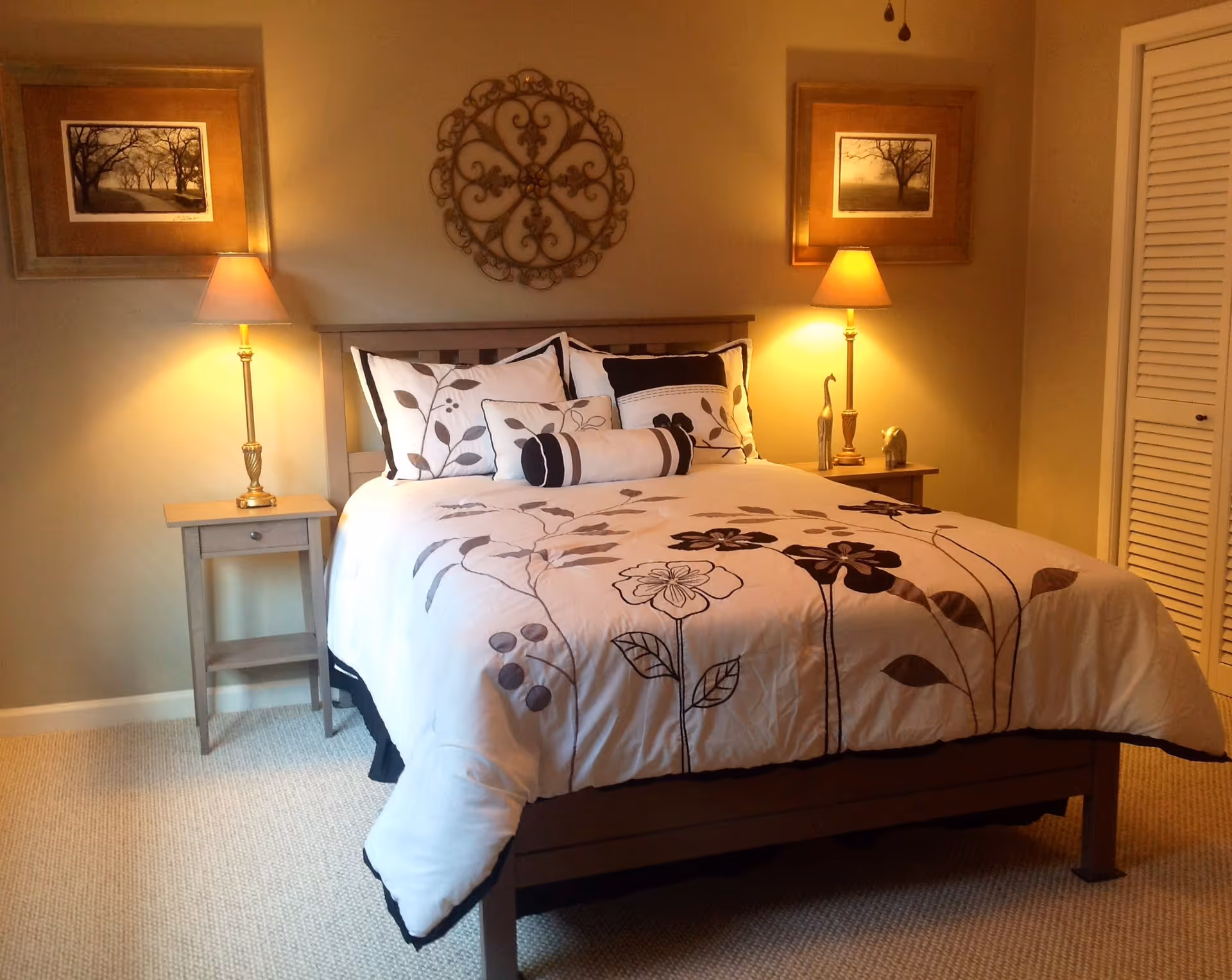 A warmly lit bedroom featuring a neatly made bed with a floral comforter, two bedside tables with lamps, and wall art above the headboard.