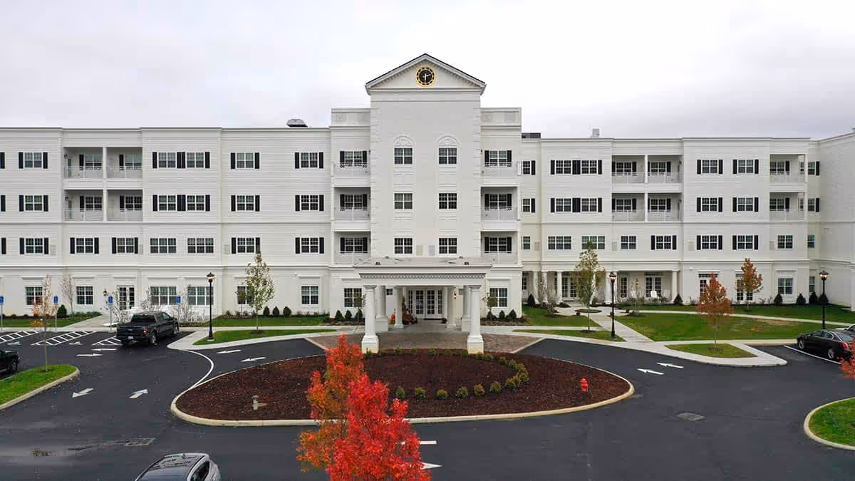 White four-story senior living building with a covered entrance, circular driveway, and landscaped grounds under an overcast sky.