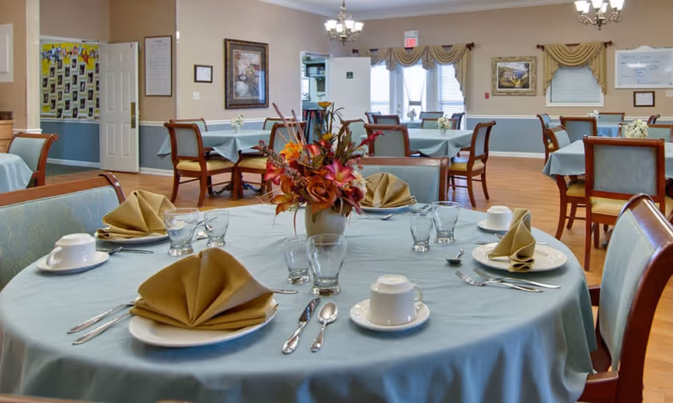 A dining room in a senior living facility with round tables covered in light blue tablecloths. Each table is set with plates, cups, utensils, and folded beige napkins. A floral centerpiece is on the nearest table. The room has wooden chairs with cushioned seats, framed artwork on the walls, and large windows with drapes allowing natural light in.