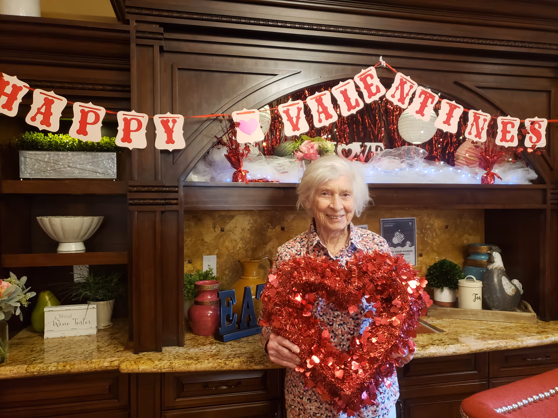 An elderly woman with white hair stands smiling in front of a wooden cabinet decorated for Valentine's Day. She is holding a large red heart-shaped decoration made of tinsel and small heart shapes. Above her, a banner reads 'HAPPY VALENTINES' with red letters on white background. The cabinet behind her has various decorative items including plants, vases, and a sign that says 'EAT'.