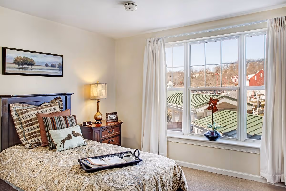 A cozy bedroom with a single bed featuring patterned bedding and multiple pillows. A wooden nightstand with a lamp, a small framed photo, and a clock sits beside the bed. A large window with white curtains offers a view of rooftops and trees outside. A small plant in a blue pot is placed on the window sill. A landscape painting hangs on the wall above the bed.