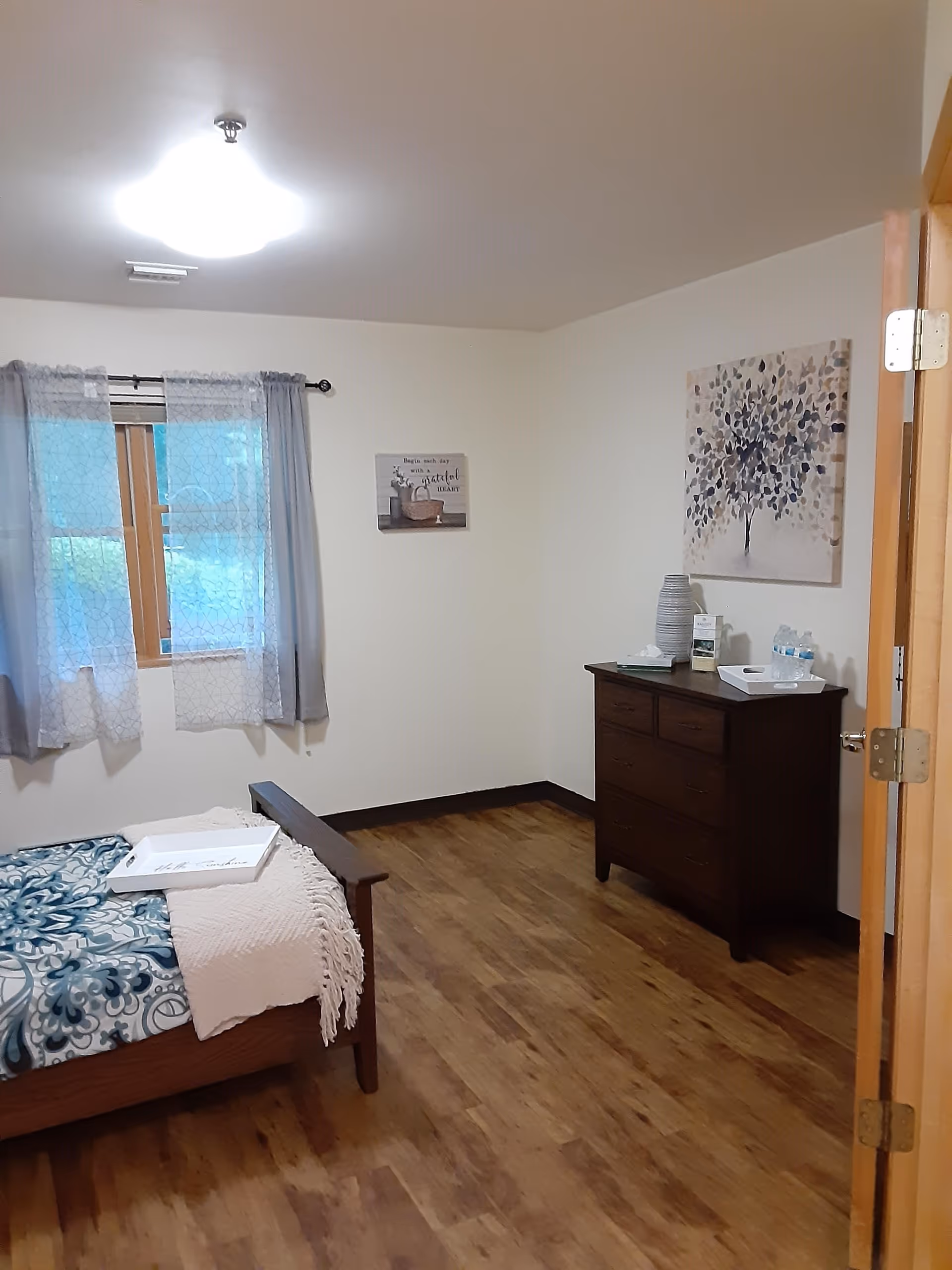 A simple bedroom with wooden flooring, a bed with a blue and white patterned blanket and a white throw, a wooden dresser with decorative items and bottled water on top, two paintings on the walls, and a window with sheer gray curtains.