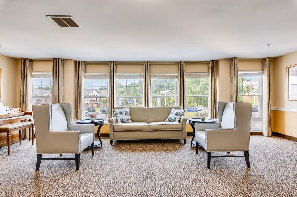A bright and spacious living room area with a beige sofa and two white armchairs arranged around two small black side tables with small flower pots. Large windows with beige curtains let in natural light and provide a view of trees and buildings outside. A wooden piano is visible on the left side of the room.