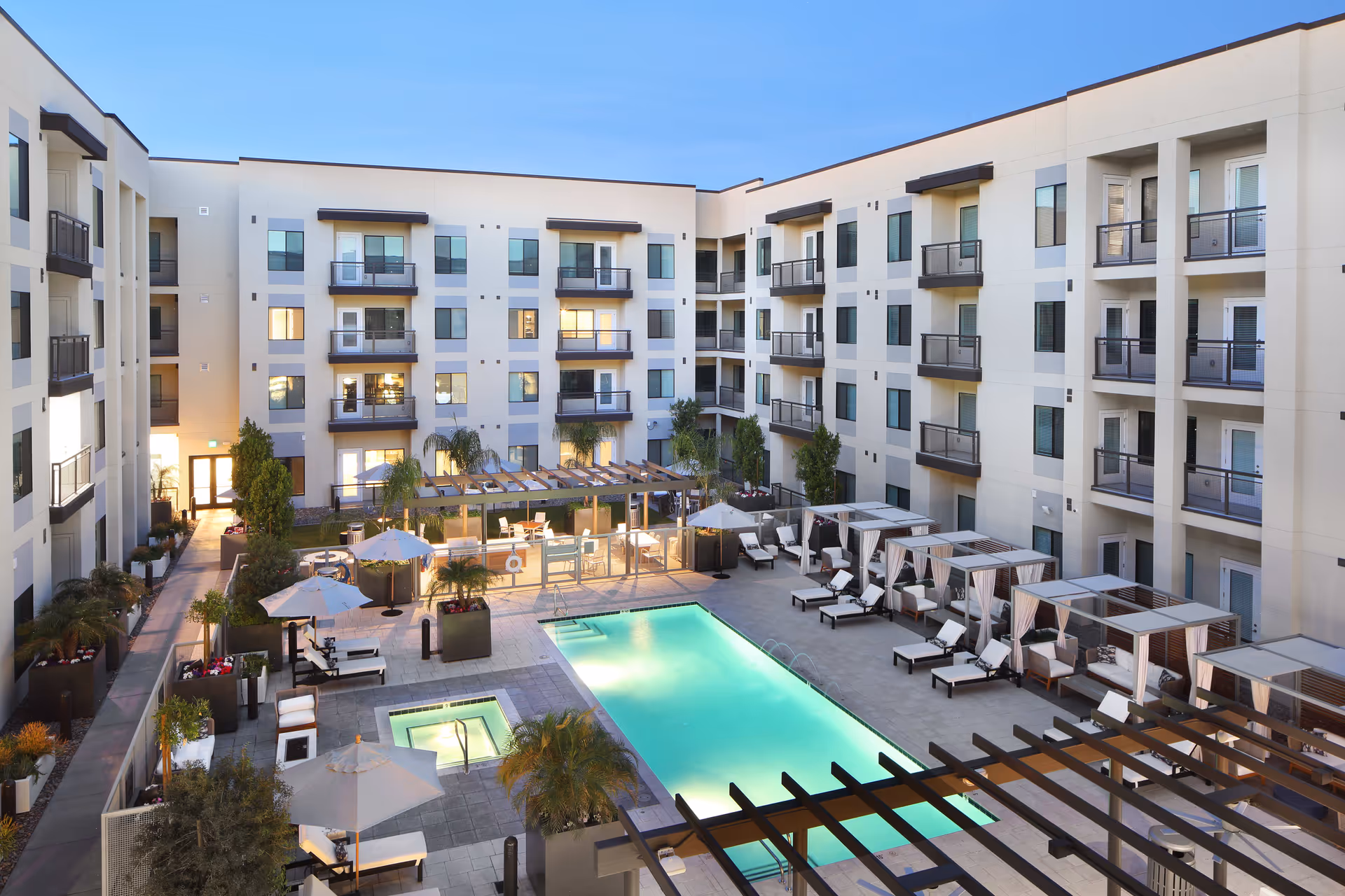 Outdoor courtyard area of a senior living facility with a swimming pool and a hot tub surrounded by lounge chairs, umbrellas, cabanas, and potted plants. The courtyard is enclosed by a four-story building with balconies and windows. The sky is clear and blue.