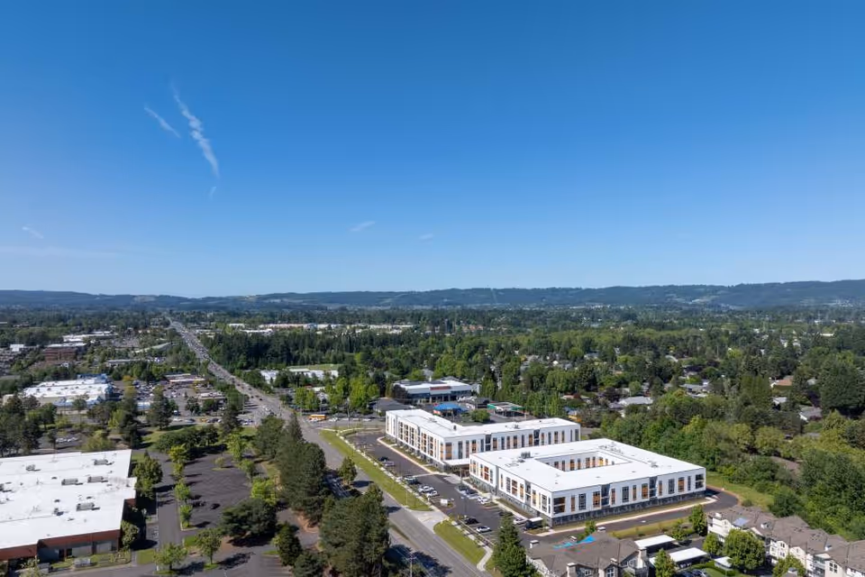Aerial view of a suburban area featuring a large, modern senior living facility with white roofs and multiple parking lots surrounded by greenery and residential neighborhoods under a clear blue sky.