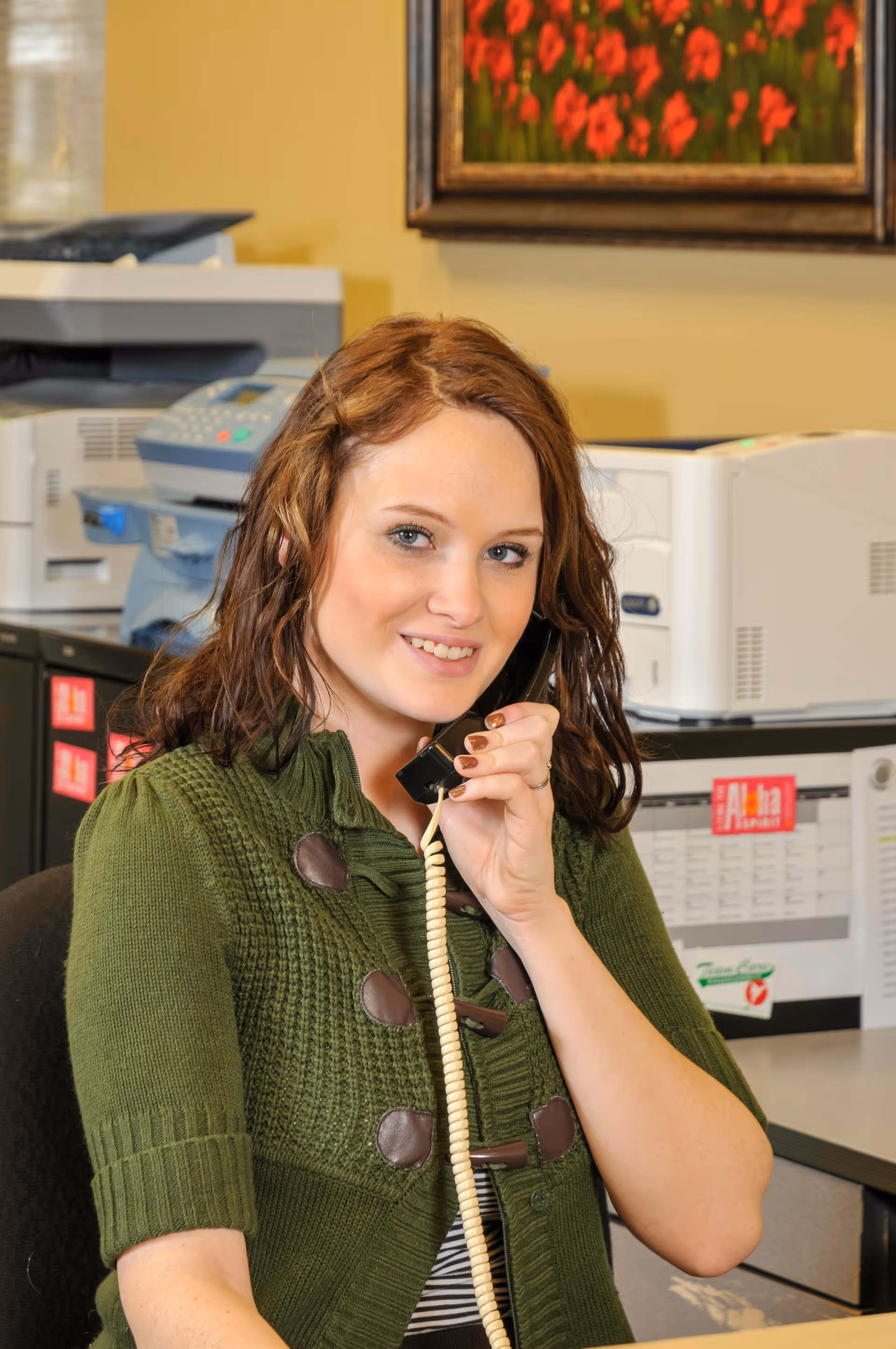 A woman with brown hair wearing a green sweater is sitting at a desk and holding a telephone receiver to her ear. Behind her are office machines including a printer and copier, and a framed painting with red flowers on the wall.