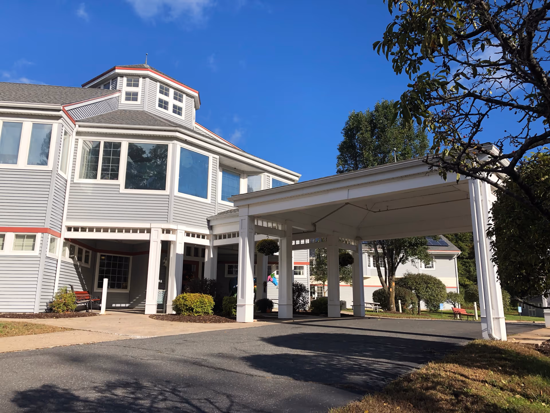 Exterior view of a senior living facility building with gray siding and white trim under a clear blue sky. The building features multiple windows and a covered entrance supported by white columns. There are trees and bushes around the building and a paved driveway leading to the entrance.