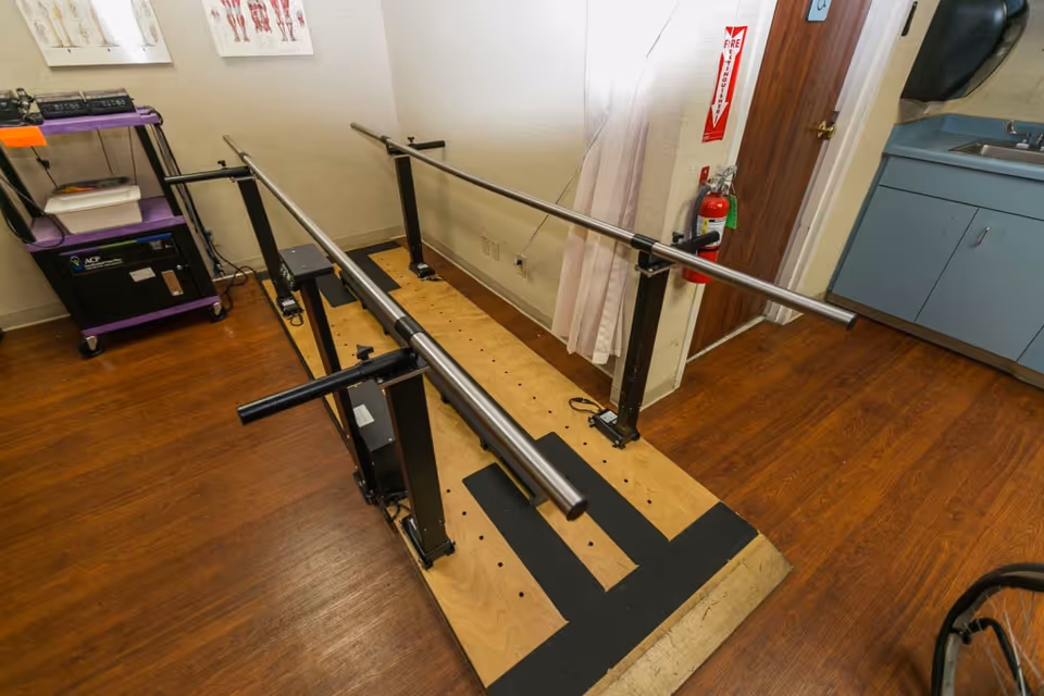 A physical therapy room with parallel bars mounted on a wooden platform for walking exercises, a purple medical cart with equipment, anatomical charts on the wall, a fire extinguisher, and a blue sink cabinet.