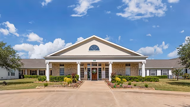 Front entrance of a single-story senior living facility with white columns, stone facade, landscaped beds, and a circular driveway under a blue sky.