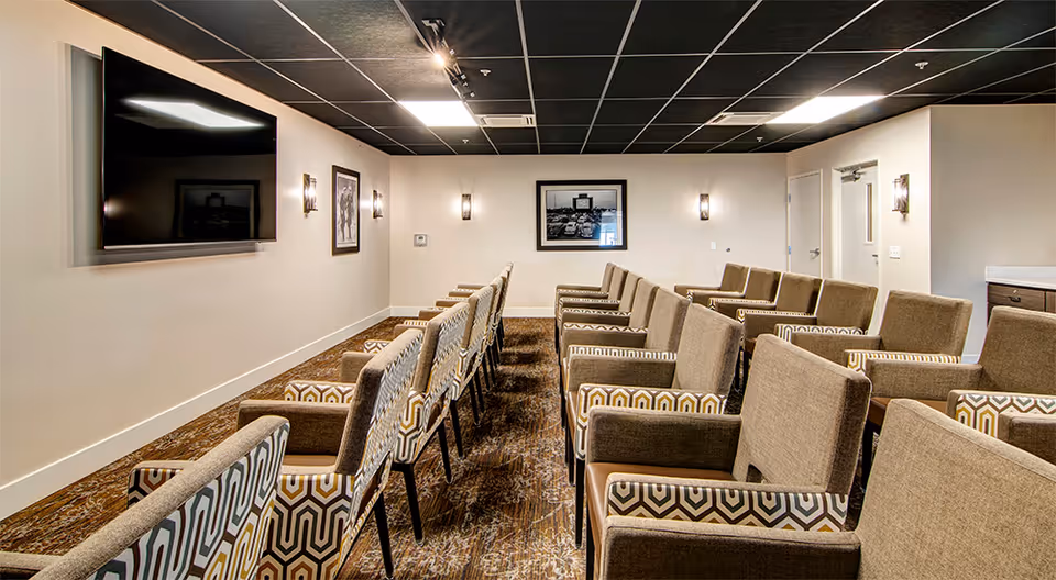 A conference or meeting room with multiple rows of cushioned chairs facing a large flat-screen TV mounted on the wall. The room has beige walls, framed black and white photographs, wall sconces for lighting, and a patterned carpet. There is a door and a small counter area visible on the right side.