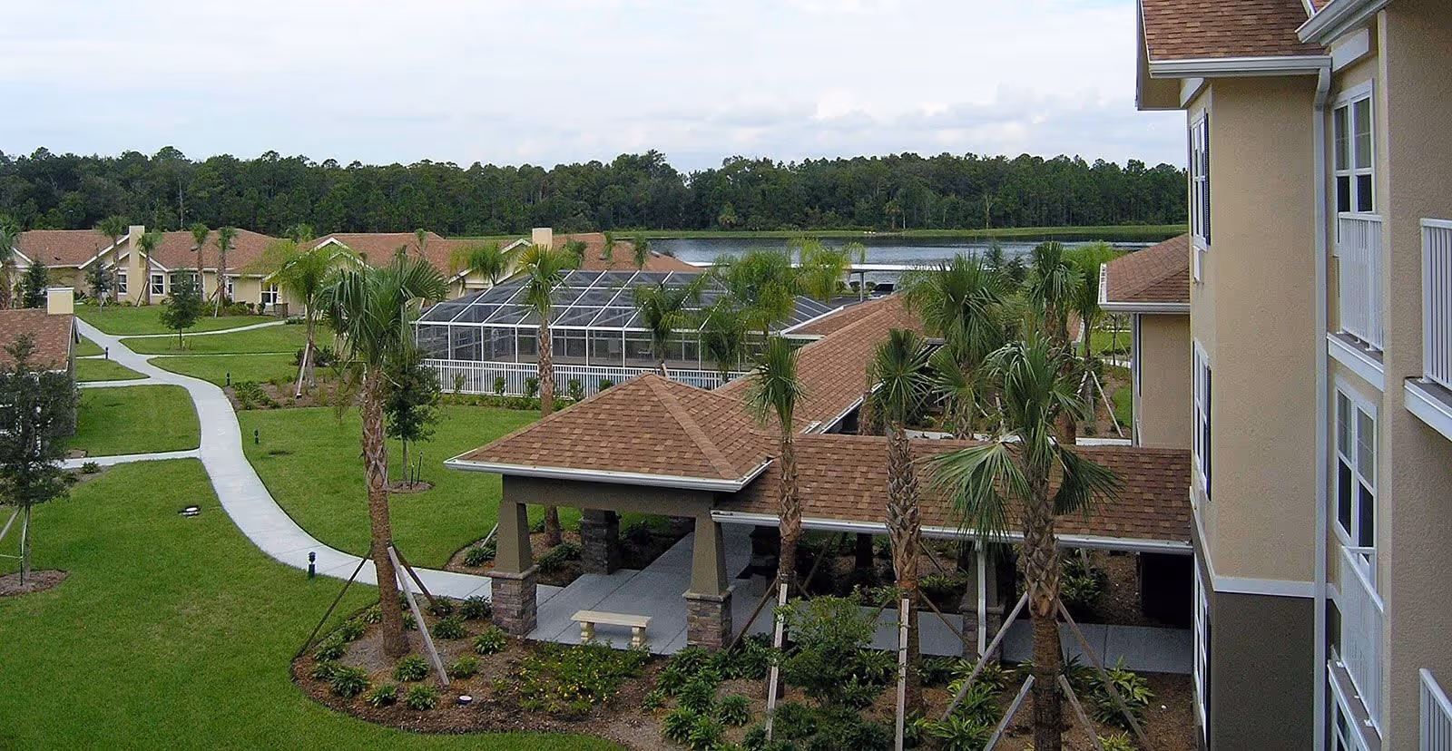 View of a senior living facility with beige buildings, palm trees, green lawns, paved walkways, and a screened-in pool area, with a wooded area and water body in the background.