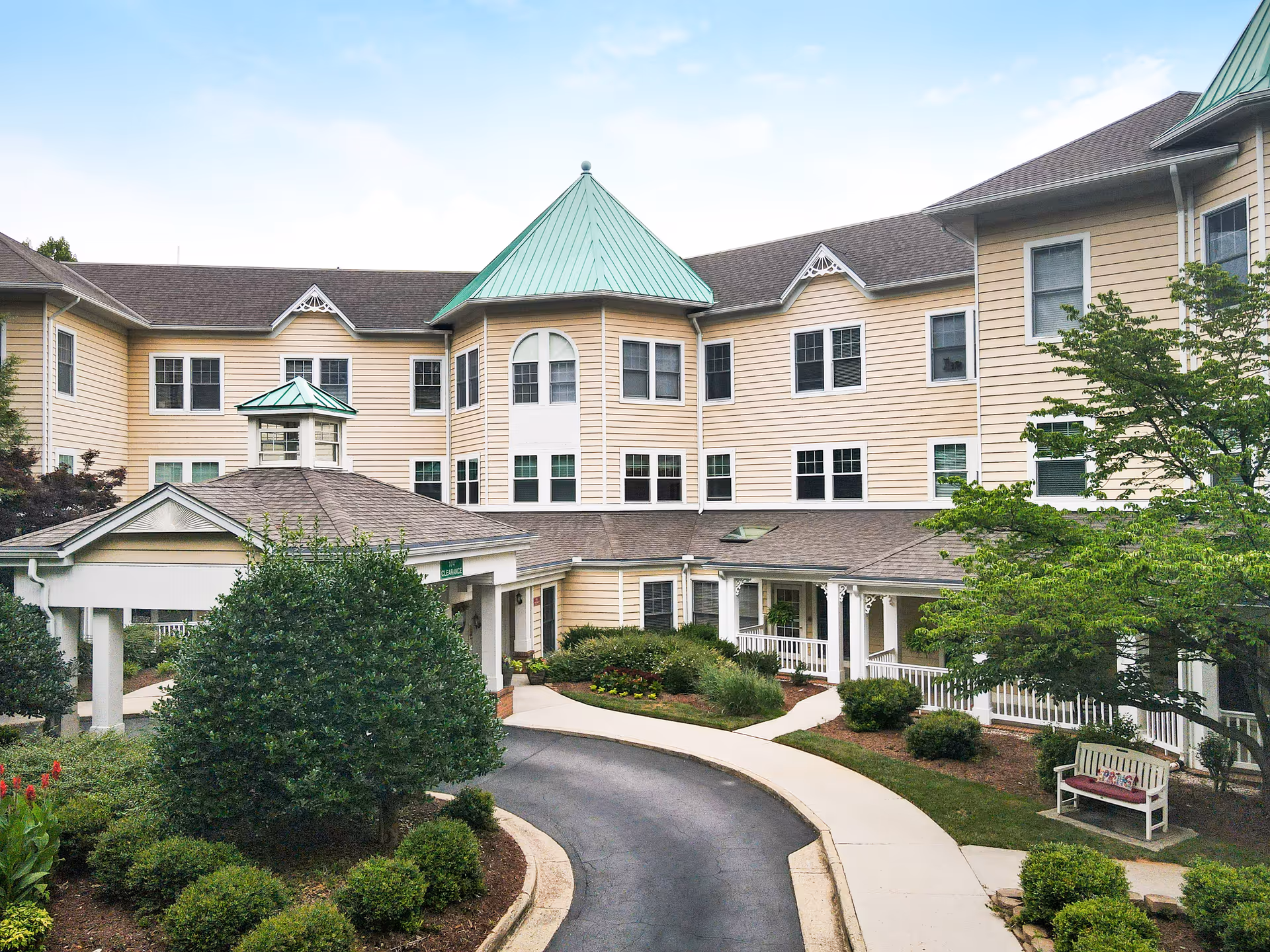 Front entrance of a three-story senior living building with a covered porte-cochere, circular driveway, and landscaped shrubs.
