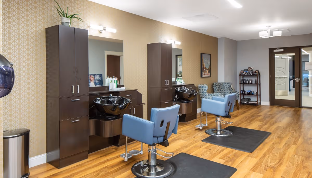 Interior view of a salon area in a senior living facility with two blue salon chairs in front of black hair washing sinks and dark wood cabinetry. The room has wood flooring, patterned wallpaper on one wall, two armchairs in the background, and double glass doors at the far end.