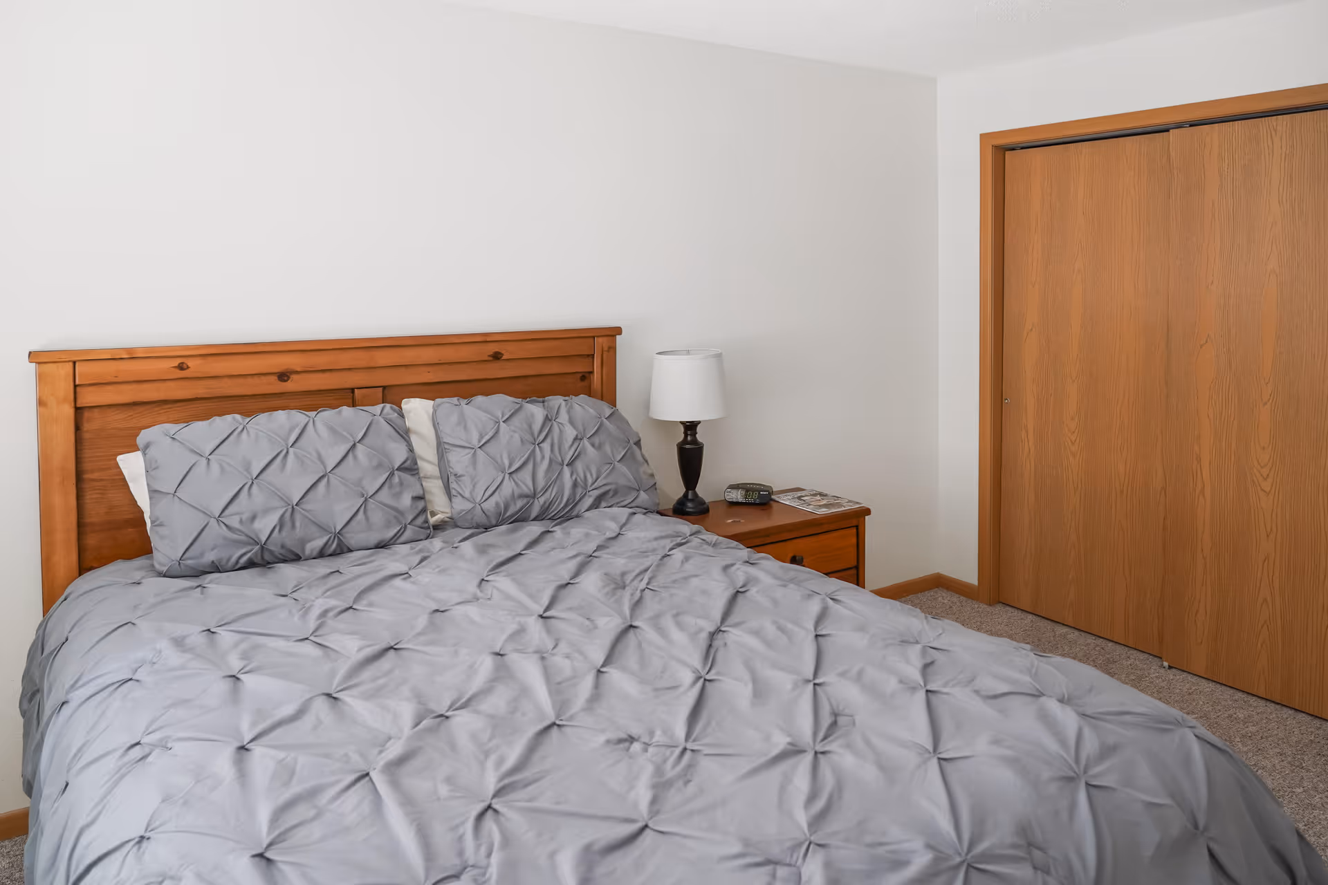 A bedroom with a wooden headboard bed dressed in gray bedding, a nightstand with a lamp and alarm clock, and sliding wooden closet doors.