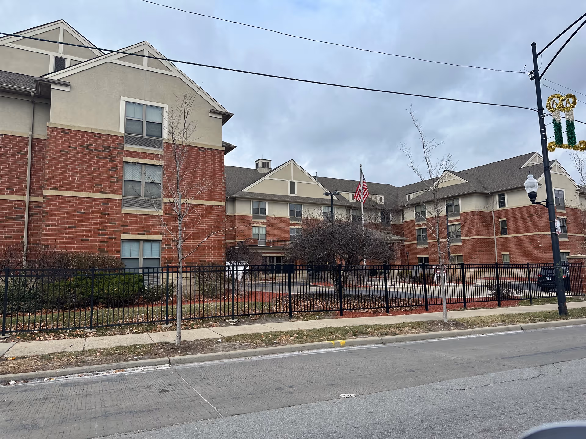 Exterior view of a multi-story senior living facility building with red brick and beige siding. The building is surrounded by a black metal fence, leafless trees, and landscaping with bushes and mulch. An American flag is displayed on a flagpole near the entrance. The sky is overcast.