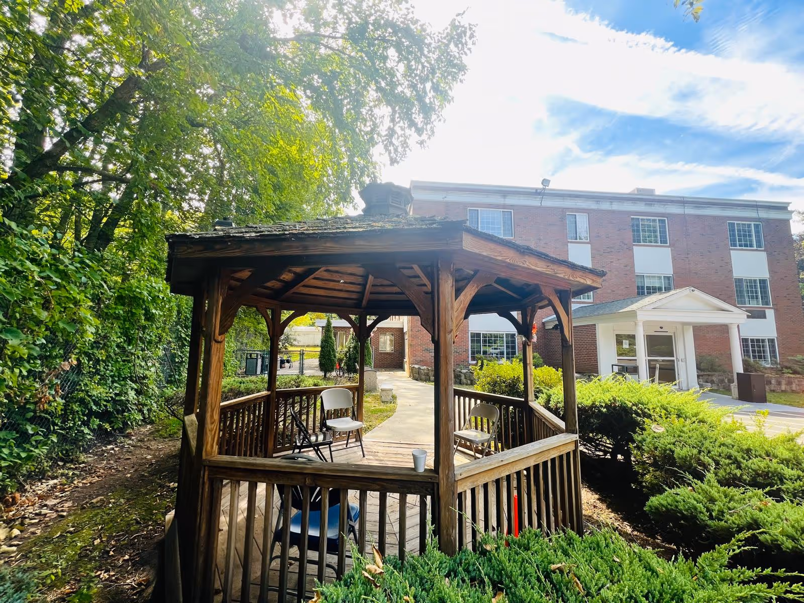 Wooden gazebo with chairs in a landscaped outdoor area in front of a brick senior living building.