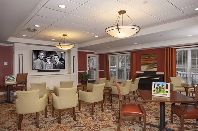 A senior living facility common area with beige armchairs arranged in rows facing a wall-mounted flat screen TV showing a black and white movie. The room has patterned carpet, red walls with white trim, large windows with red curtains, a piano, and several tables with chairs. Ceiling lights illuminate the space.
