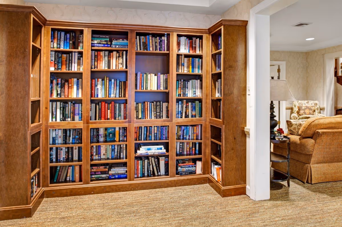 A cozy interior corner featuring a large wooden bookshelf filled with various books. To the right, there is a glimpse of a living area with a brown upholstered sofa, a floral armchair, a side table with a lamp, and beige patterned wallpaper.
