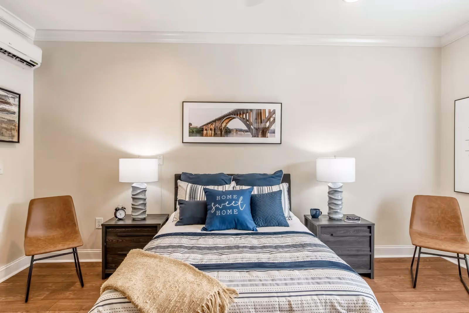 A neatly made bed with striped bedding and multiple pillows, including a blue pillow with the words 'Home Sweet Home'. The bed is flanked by two dark wooden nightstands, each with a modern lamp and small decorative items. Two brown chairs are placed on either side of the nightstands. The walls are light-colored and decorated with framed pictures, including a large photo of a bridge above the bed.