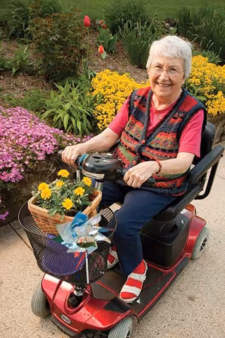 An elderly woman with white hair and glasses is smiling while sitting on a red mobility scooter. She is wearing a colorful patterned vest over a pink shirt and red sandals. The scooter has a basket with yellow flowers and a decorative pinwheel attached. She is outdoors on a paved path with vibrant flower beds featuring yellow, pink, and red flowers in the background.