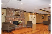 Spacious lounge area with hardwood floors, a brick fireplace, upholstered chairs and framed artwork.