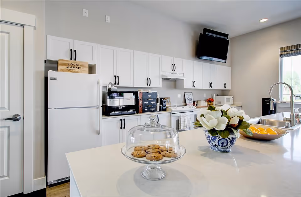 A modern kitchen with white cabinets, a white refrigerator, a stove, microwave, and coffee machines. On the white countertop island in the foreground, there is a glass cake stand with cookies, a blue and white vase with white flowers, and a bowl of lemons. A flat-screen TV is mounted on the wall above the stove, and a window with a striped blind lets in natural light.