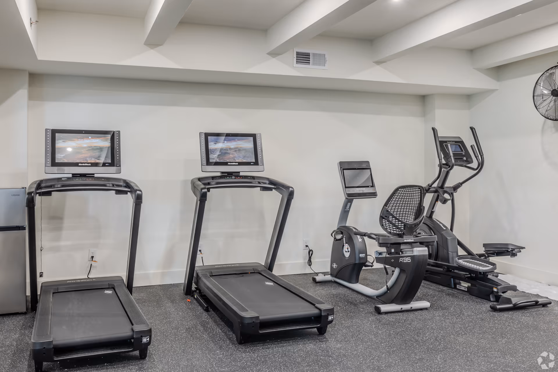 Indoor fitness room with two treadmills, a recumbent exercise bike, and an elliptical machine against a white wall with a ceiling fan mounted on the right side.