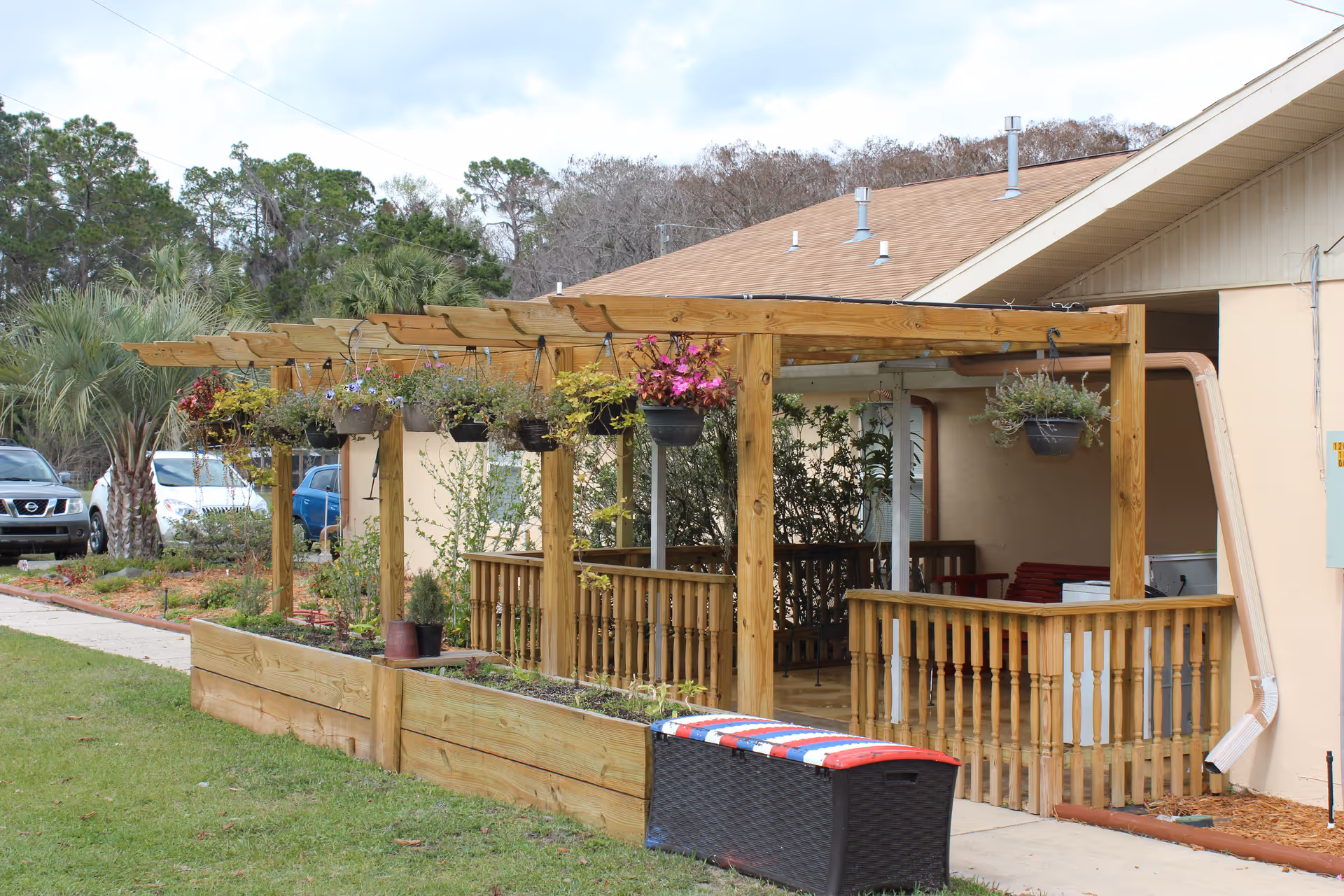 Outdoor patio area with a wooden pergola structure supporting hanging flower pots. The patio is adjacent to a building with a beige exterior and a brown roof. There are wooden railings around the patio and a black storage bench with a red, white, and blue cushion on top. Several cars and trees are visible in the background.