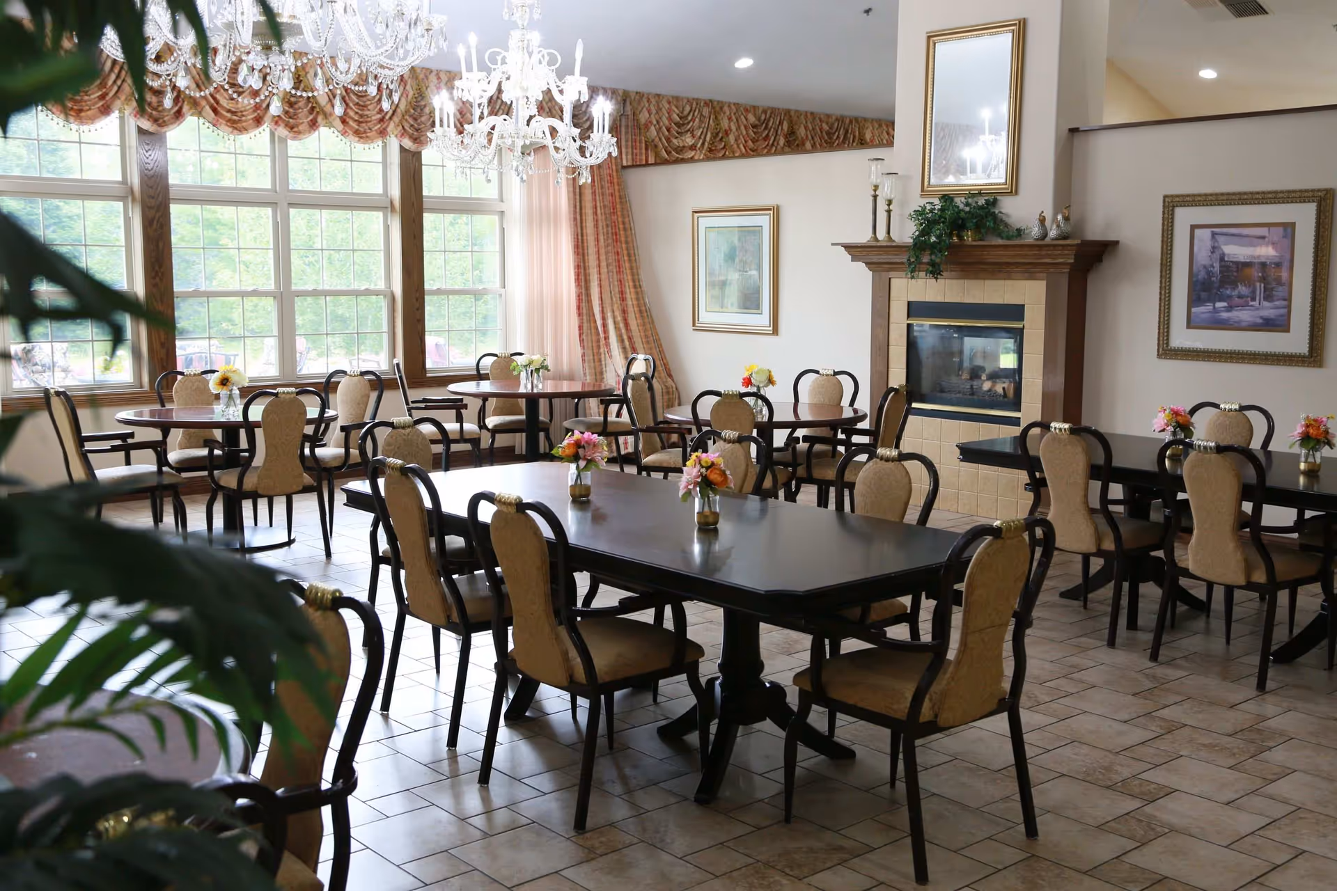A bright dining room with multiple dark wood tables and beige upholstered chairs arranged neatly. Each table has a small vase with colorful flowers. Large windows with decorative curtains allow natural light to fill the room. A fireplace with a wooden mantle and framed artwork adorn the walls, and two chandeliers hang from the ceiling.