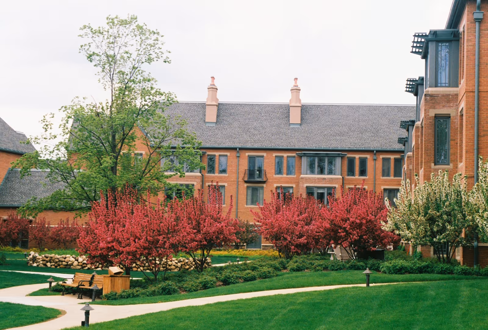 A landscaped outdoor courtyard area with green grass, walking paths, benches, and flowering bushes in front of a multi-story brick building with multiple windows and chimneys.
