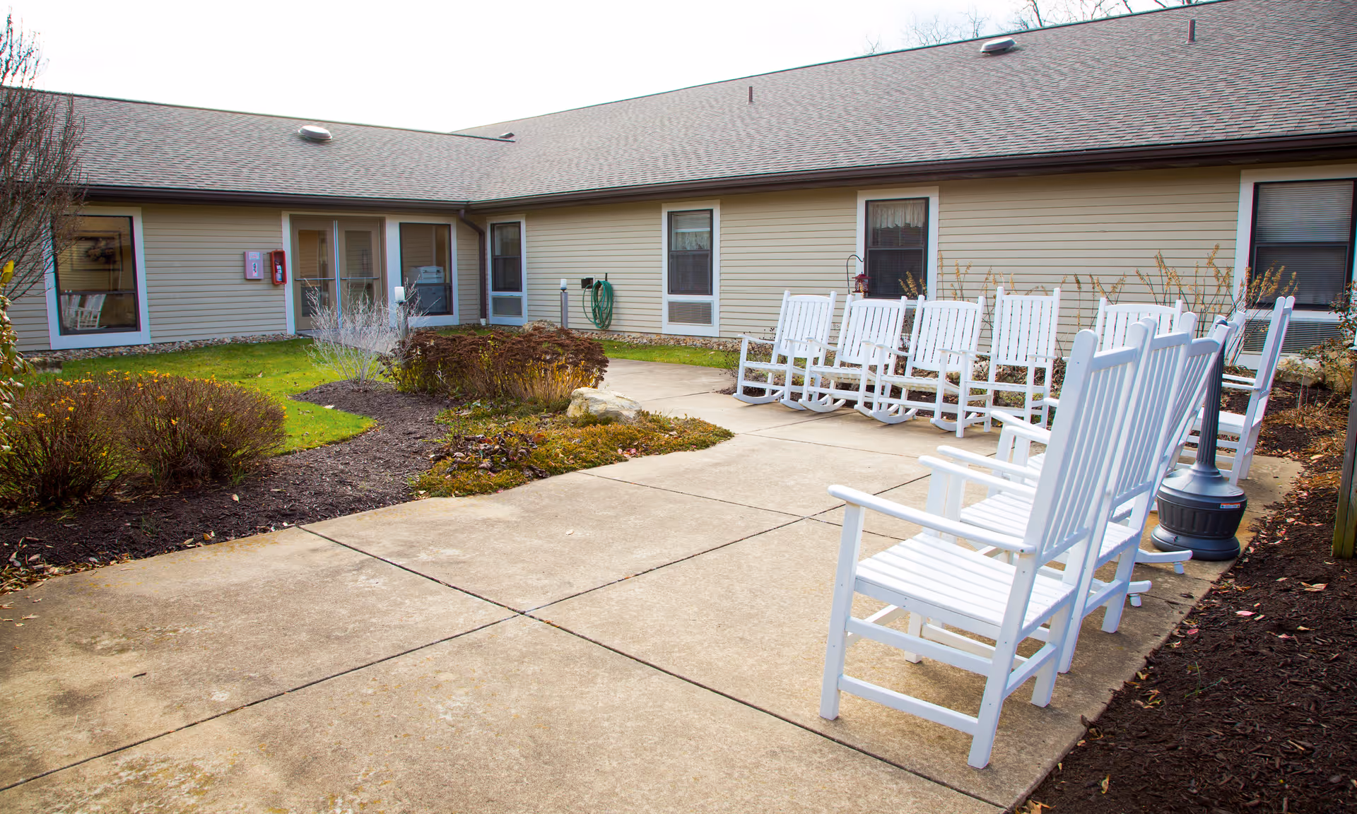 Outdoor courtyard area at Brookdale Latrobe featuring a concrete pathway, landscaped garden beds with shrubs, and a row of white wooden rocking chairs arranged along the pathway next to the building.