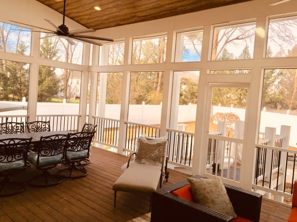A screened-in porch area with a wooden floor and ceiling, featuring a large ceiling fan. There is a black metal dining table with six matching chairs on the left side, a cushioned lounge chair in the center, and a cushioned armchair with a pillow on the right. The porch overlooks a fenced backyard with trees and shrubs visible outside.