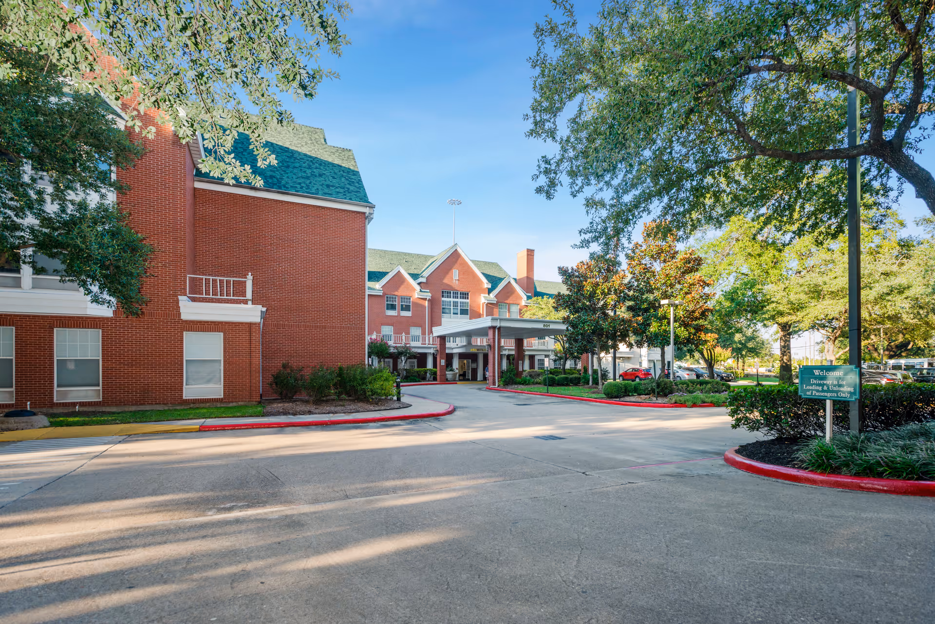 Exterior view of a senior living facility with red brick buildings, green roofs, and a covered entrance driveway. Trees and landscaped bushes surround the driveway under a clear blue sky.