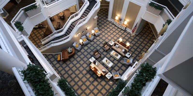 View from above of a spacious indoor common area with patterned carpet, multiple seating arrangements including sofas and armchairs with cushions, side tables with lamps, a piano, and staircases with white railings. Green plants are placed along the railings and an American flag is visible in the corner.