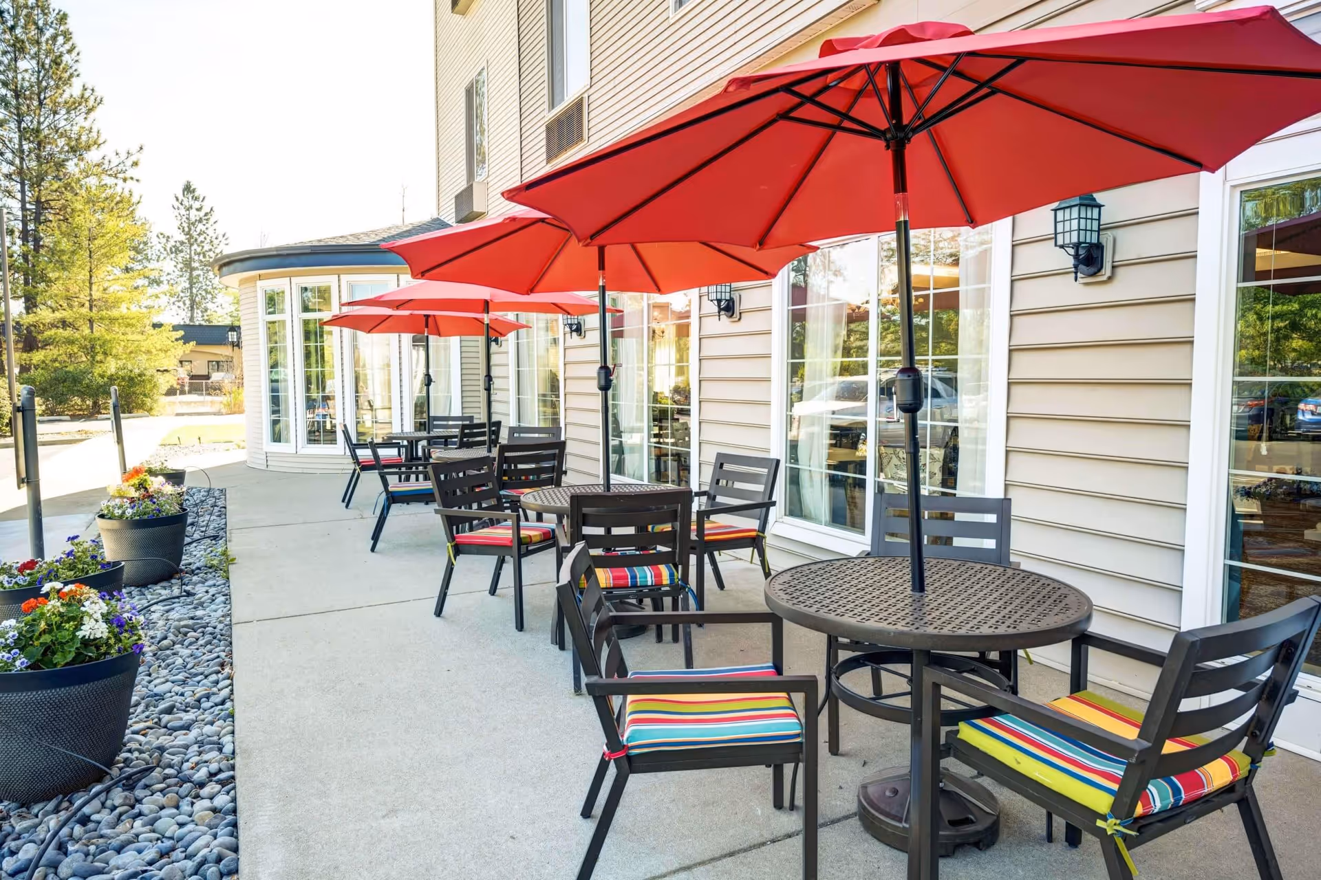 Outdoor patio area with round metal tables and chairs that have colorful striped cushions. Each table has a large red umbrella providing shade. The patio is adjacent to a beige building with large windows and is decorated with flower pots containing colorful flowers.