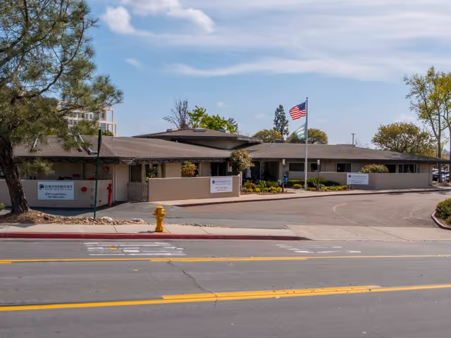 Front view of Grossmont Post Acute Care single-story building with flags, entrance signage, landscaping, and a fire hydrant at the curb.