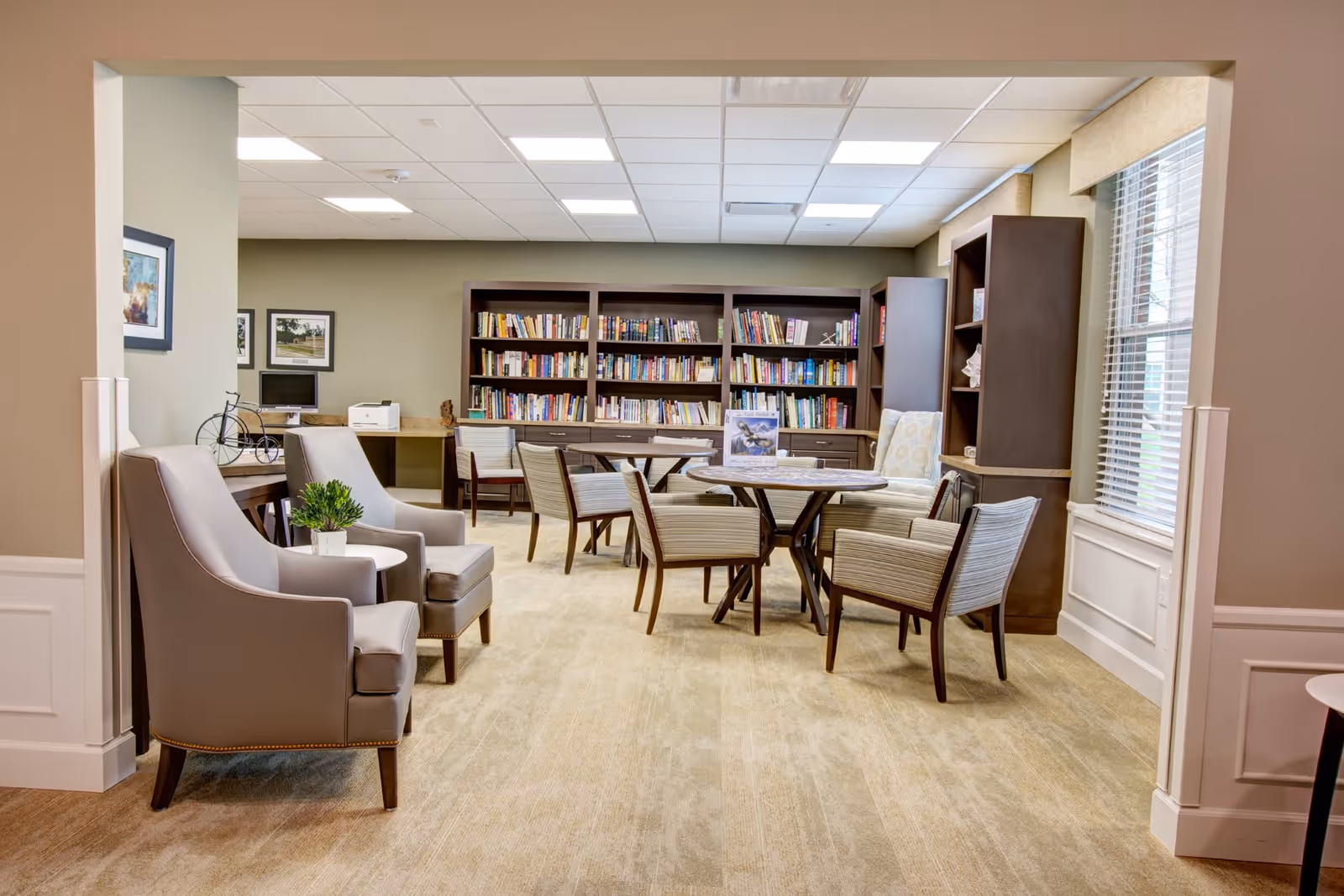 A cozy reading and sitting area in a senior living facility with beige carpet, two gray armchairs with a small round table between them, a round table surrounded by six striped chairs, and a large bookshelf filled with books against the back wall. There is a window with blinds on the right side and framed pictures on the left wall.